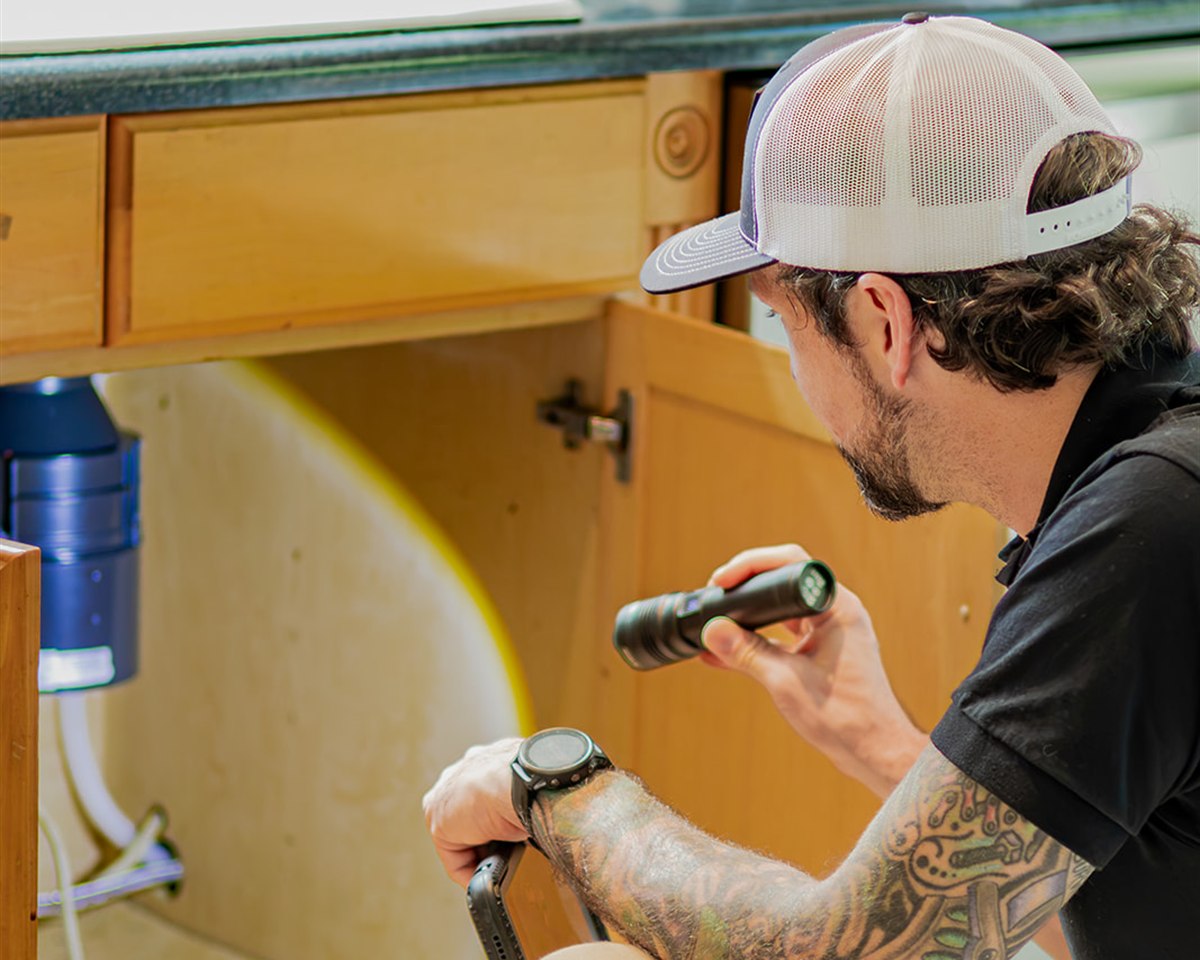 Frame inspector checking plumbing under a kitchen sink with a flashlight